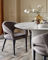 Elegant round marble table with curved grey velvet chairs, accented by a vase of flowers and decorative tableware.