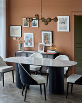 Modern dining table with oval marble top, black fluted base, and upholstered chairs in two-tone fabric. Gallery wall and pendant light.
